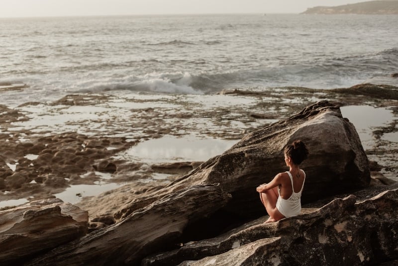 woman in white swimsuit sitting on rock looking at sea