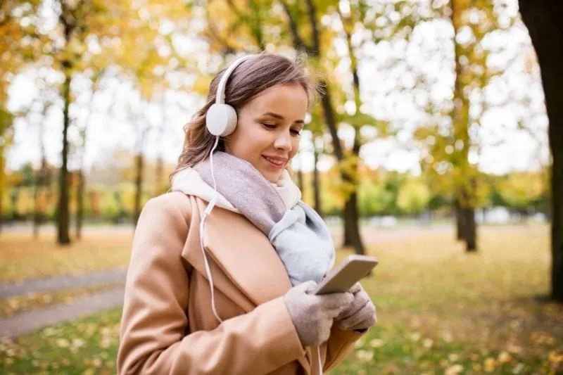 woman with a smartphone and headphone smiling and standing in the park and smiling
