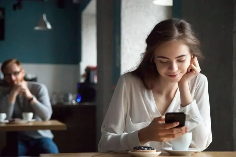 young curious guy looking a cute woman in a nearby table at a cafe looking smiling at her smartphone