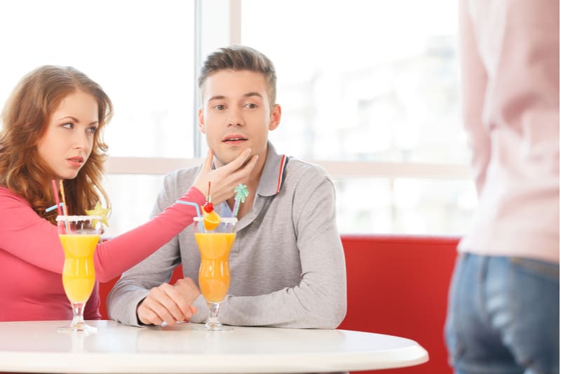 young man looking at lady passing by while being stopped by the woman sitting beside him by the table