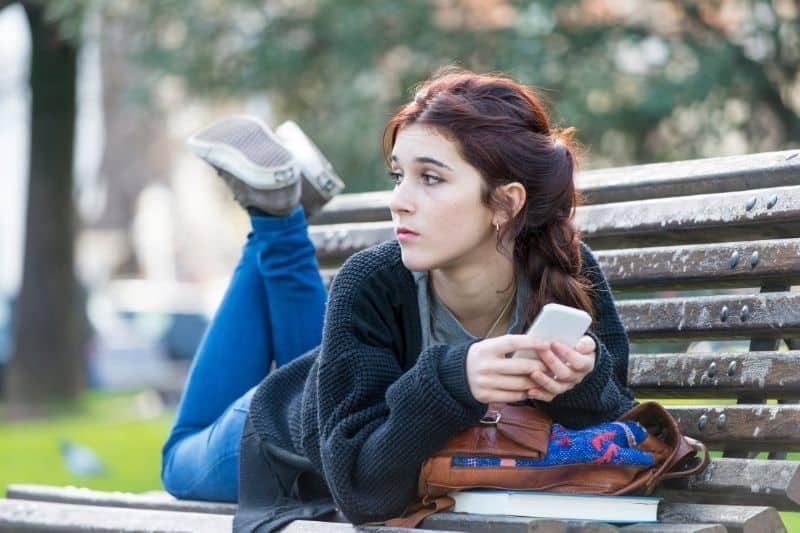 young student adolescent girl waiting at the school's bench with her cellphone and books on the bench