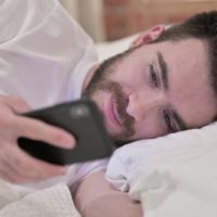 man reading text message in his cellphone while lying by his side on bed