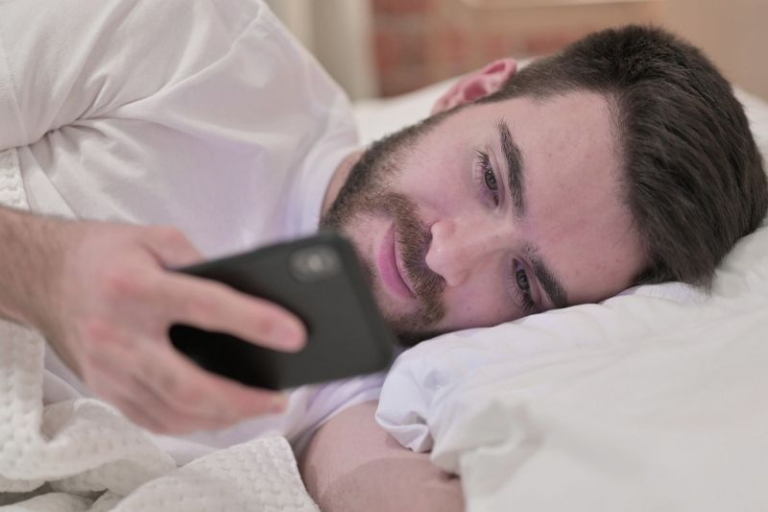 homem a ler uma mensagem de texto no seu telemóvel enquanto está deitado na cama