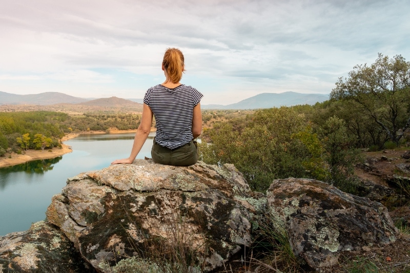 woman in striped t-shirt sitting on rock looking at lake