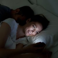 donna che sorride e manda messaggi al cellulare mentre è sdraiata accanto al marito che dorme