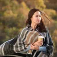 mulher enrolada num cobertor, segurando uma chávena de café, a caminhar ao ar livre