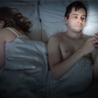 man reading mesages on his cellphone while lying in bed beside his sleeping girlfriend