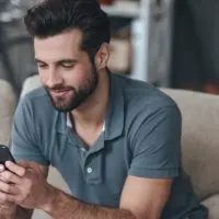 handsome man texting on phone sitting on the couch inside living room and smiling