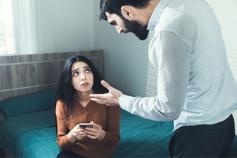 uomo arrabbiato sopra la moglie che tiene un cellulare seduto sul letto all'interno della camera da letto