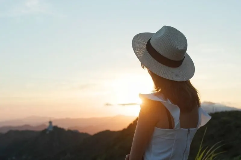 backview of a woman looking at the sunset wearing hat and white dress standing on top of a mountain
