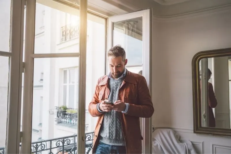 bearded man reading text from his smartphone standing near the glass door inside home