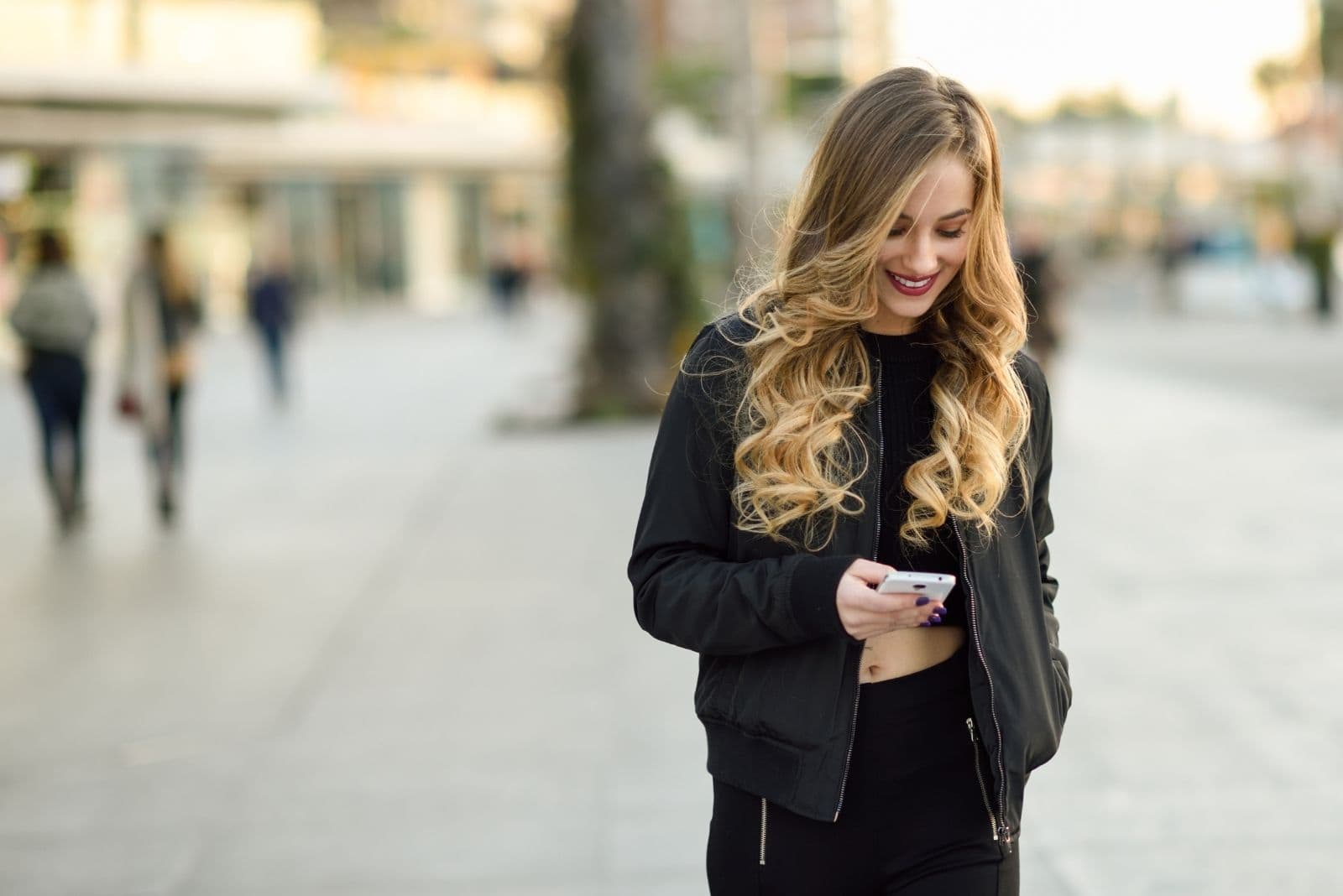 donna bionda dai capelli mossi che sorride e manda messaggi mentre cammina per strada in città