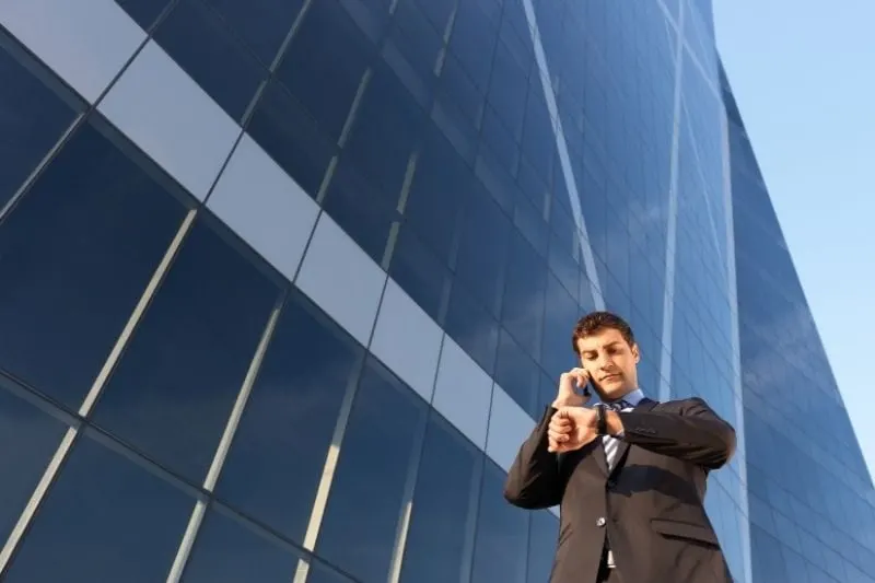 busy businessman on the phone checking his timepiece walking near a tall building phone on low angle