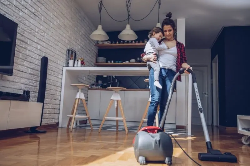 busy mother cleaning using a vacuum cleaner inside the house carrying a toddler