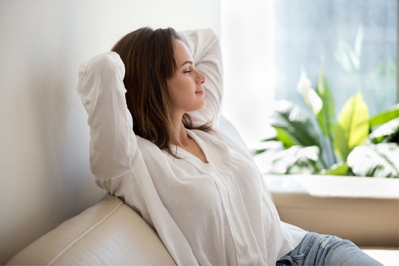 calm woman sitting on the couch in the living room closing her eyes with hands at back of her head