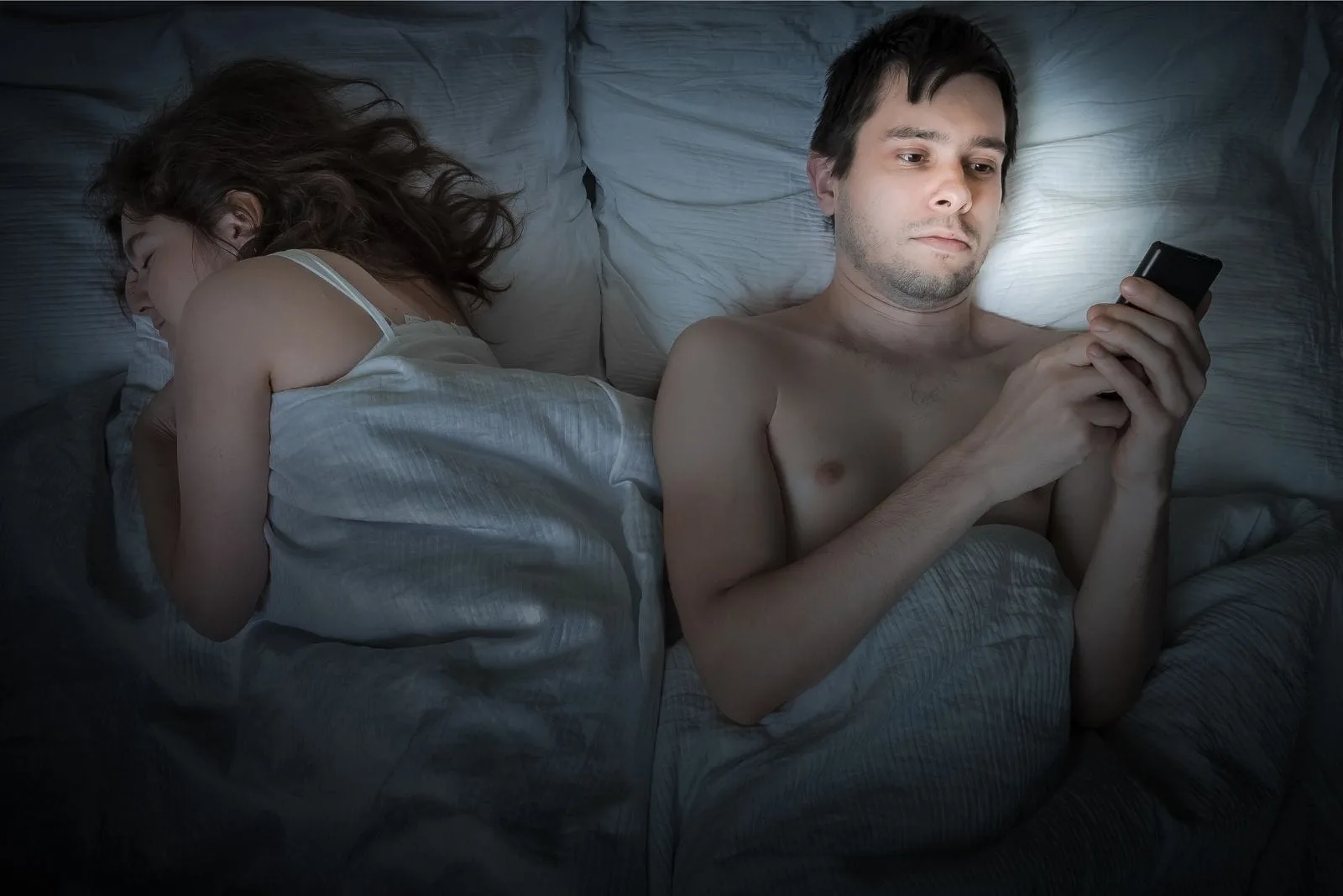 cheating man reading messages from his cellphone while lying down on bed beside his sleeping wife