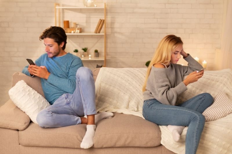 coppia che si annoia con lo smartphone e che siede ai lati opposti del divano in salotto