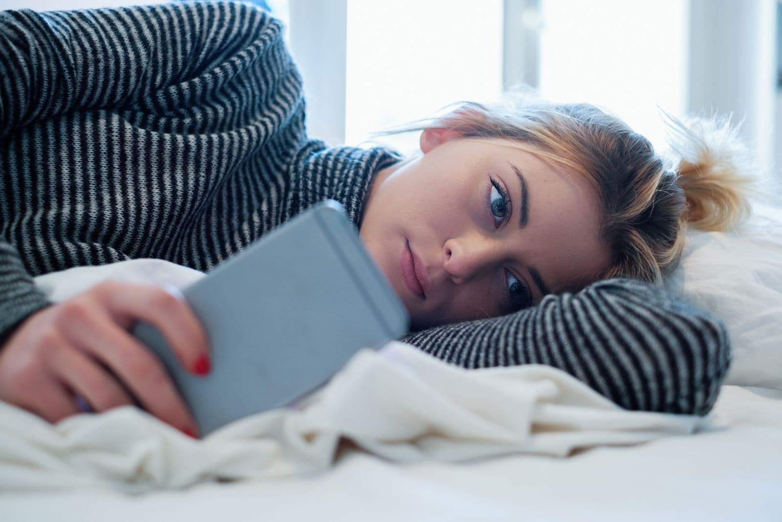 Donna delusa sdraiata a letto mentre tiene in mano il suo smartphone all'interno della camera da letto