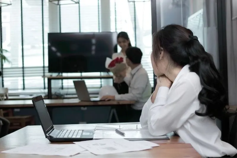 envious business woman looking at a sweet couple colleague receiving a bouquet of flower inside office