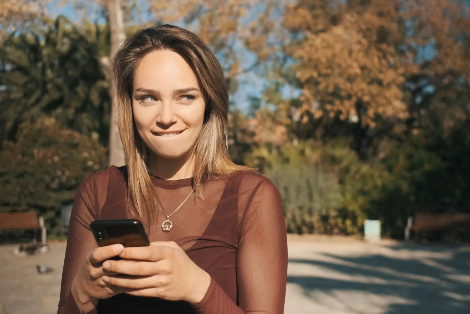 flirty woman texting biting her lip while standing outdoors and looking away