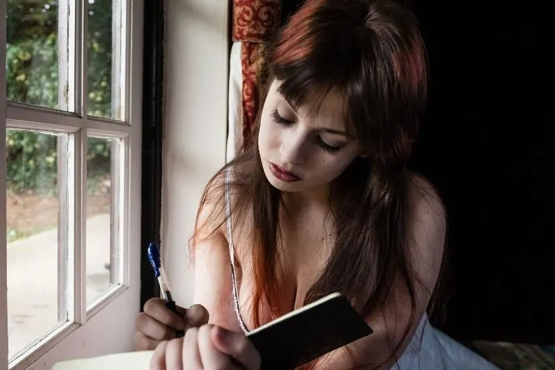 girl writing in her diary near the window of the room