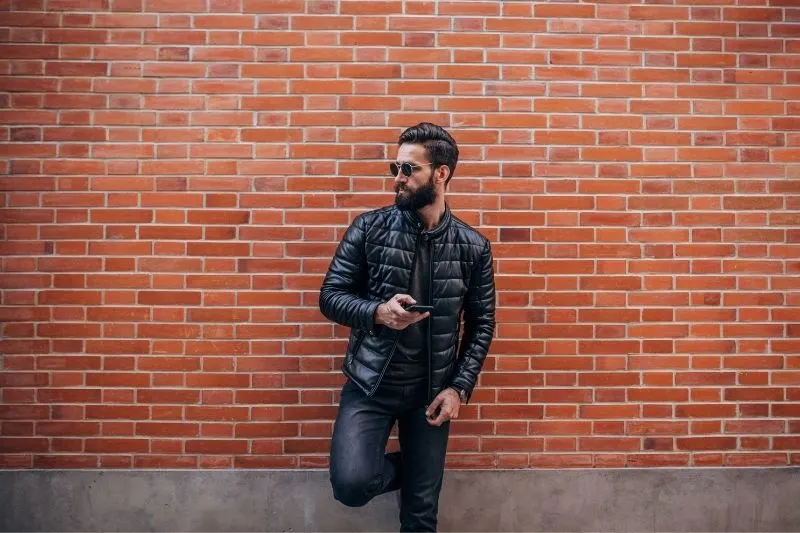 handsome bearded man texting on smartphone while leaning on the brown bricked wall
