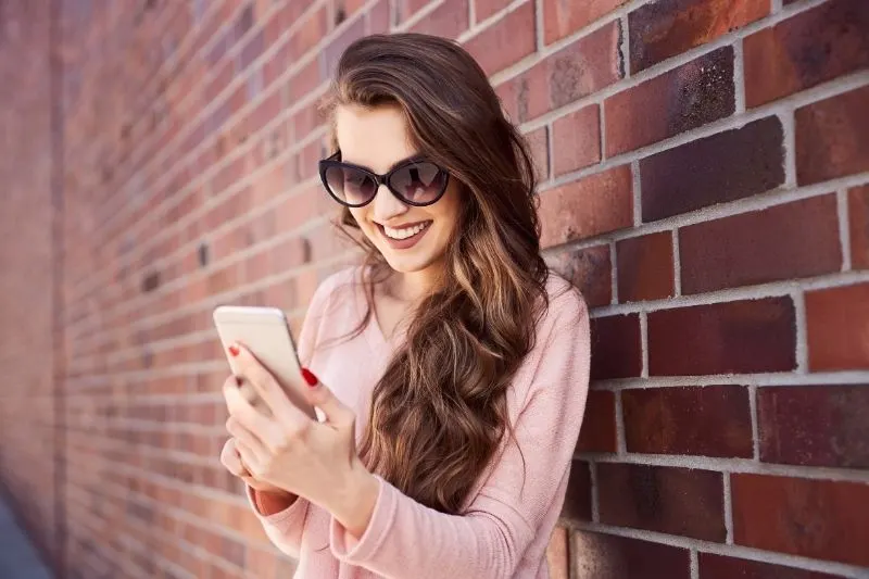 happy woman reading message on her smartphone while standing near a brick wall