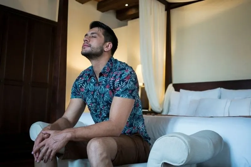 latin man sitting on the edge of the bed of the hotel closing eyes
