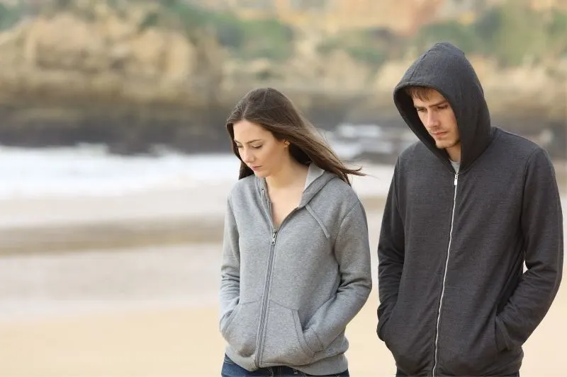 man and woman walking along the beach wearing hoodies with hands inside the pocket