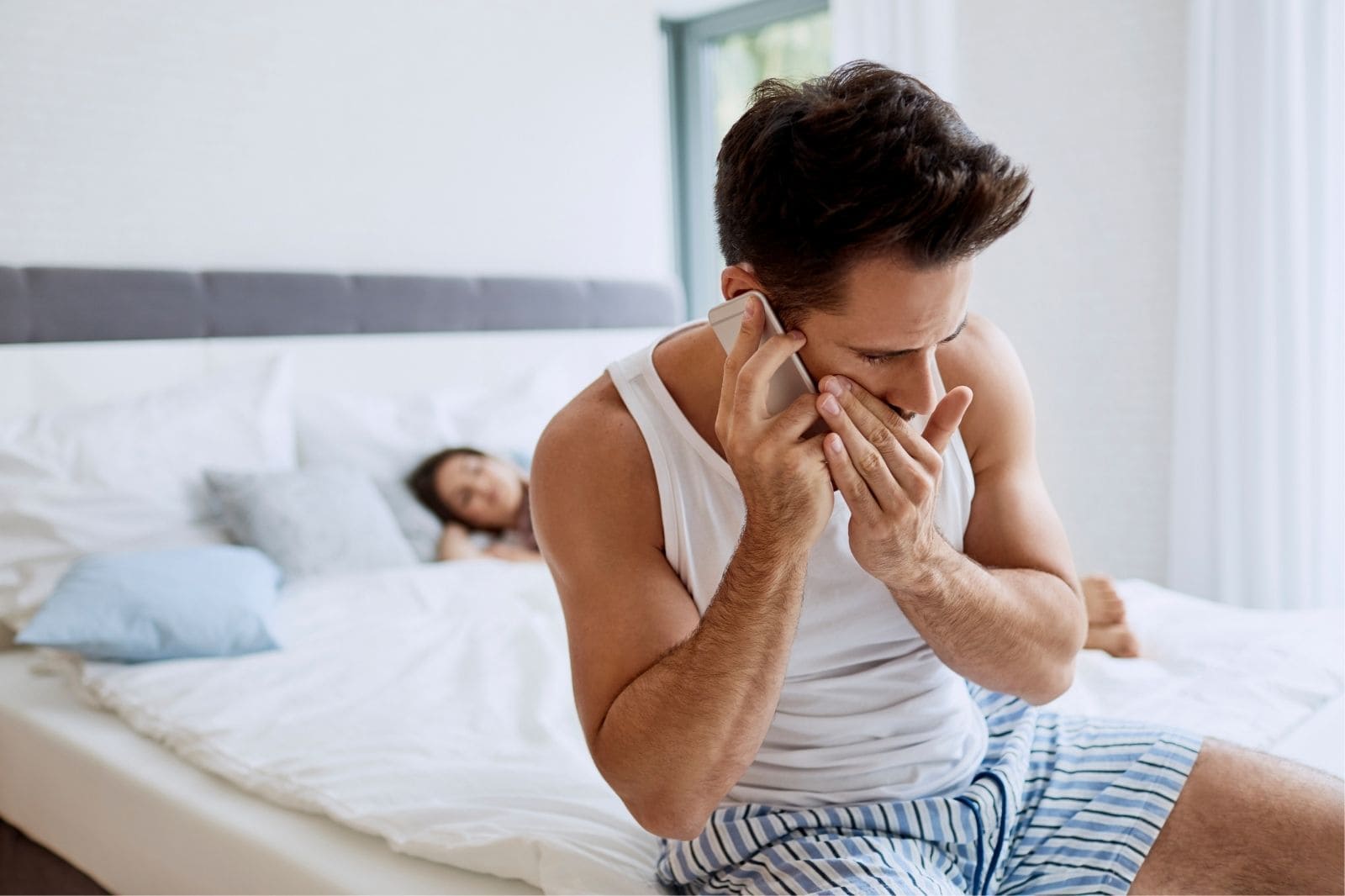 man answering phone whispering with wife sleeping on bed