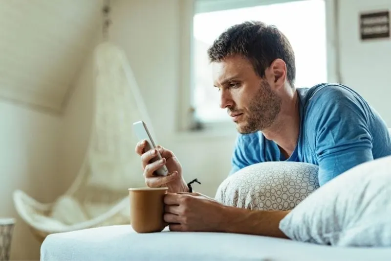 man having coffee on bed while reading his smartphone thoughtfully
