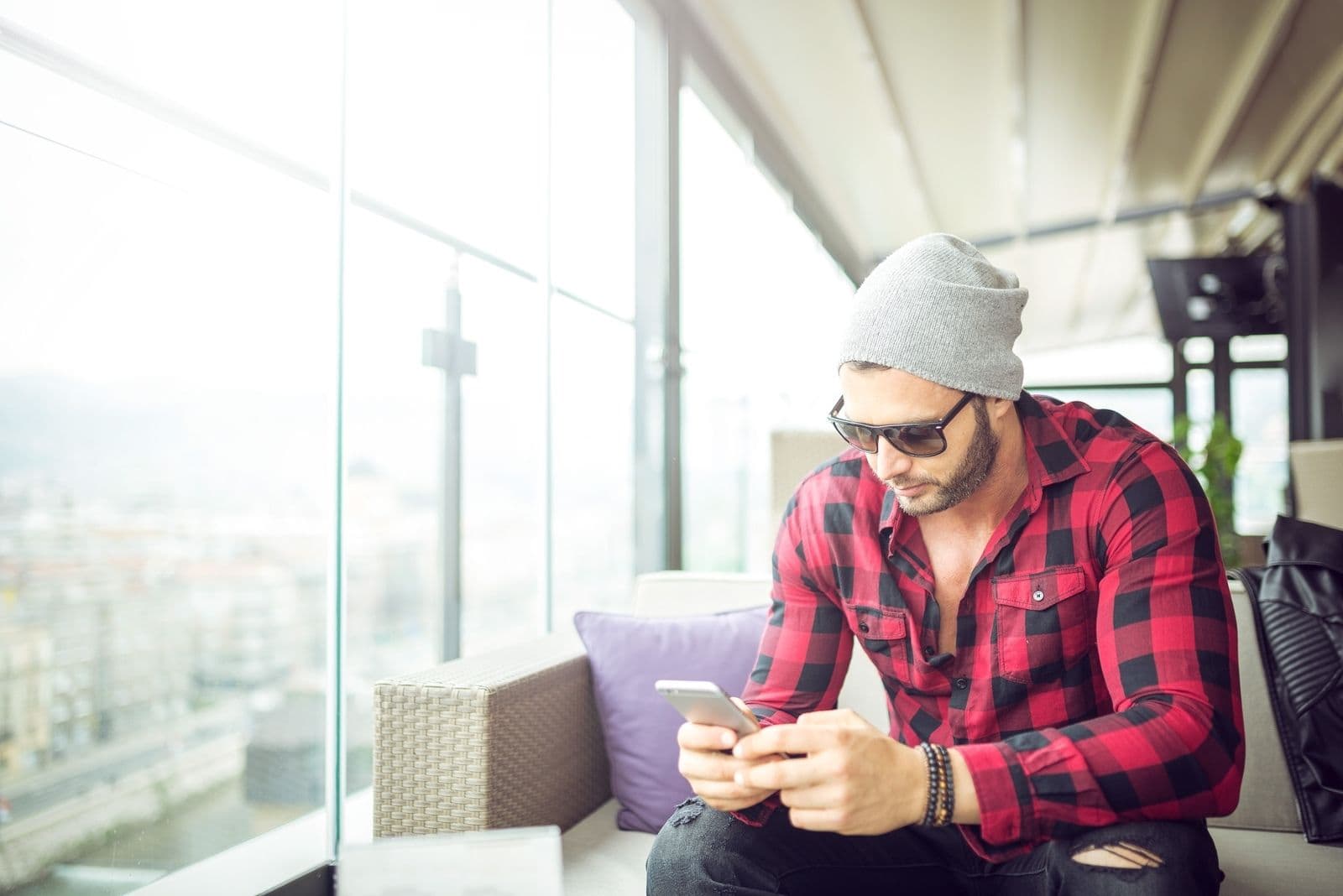 uomo con un cappellino che manda messaggi all'interno di un caffè seduto da solo vicino alla finestra e con gli occhiali da sole