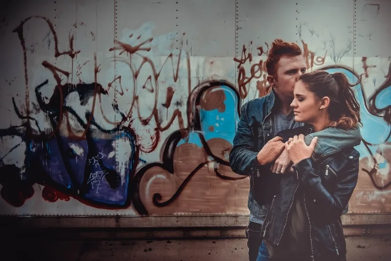 man kissing and hugging woman while standing near graffiti wall
