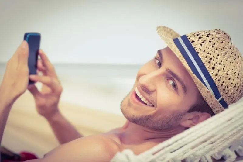 man relaxing in hammock texting on smartphone and smiling looking at the camera behind him