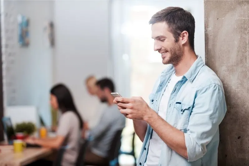 man texting and smiling while standing and leaning on a wall with colleagues working