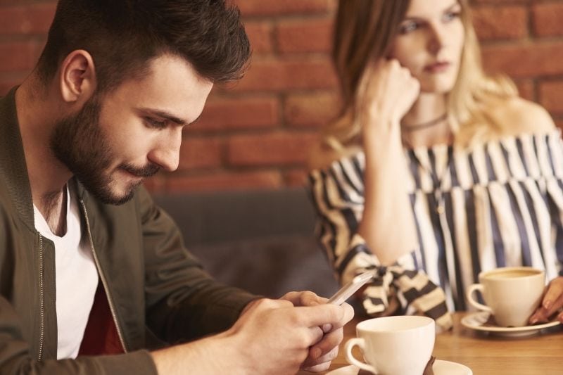 hombre escribiendo en su móvil durante una cita con una mujer con cara de aburrimiento y mirando hacia otro lado