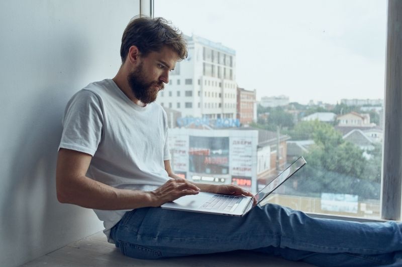 man typing on laptop squatting on the floor near the big glass window