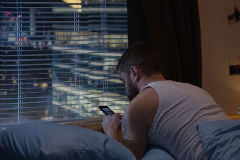 scatto medio di un uomo che utilizza il suo telefono sul letto all'interno di una stanza da un edificio alto durante la notte