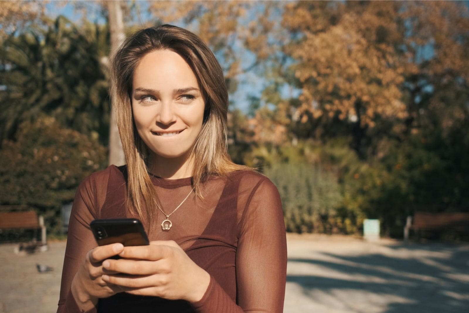 mujer traviesa sosteniendo un smartphone mordiendose el labio mientras esta de pie al aire libre