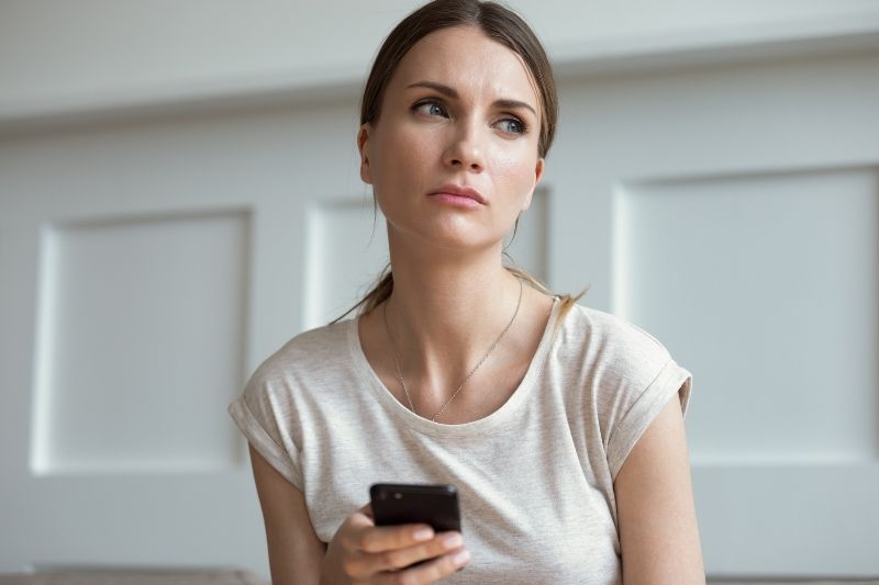 donna pensierosa sui 30 anni che tiene in mano un cellulare e indossa un top bianco all'interno della casa