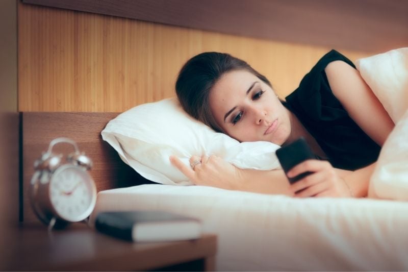 mujer triste revisando su teléfono en la cama con un despertador en la mesa