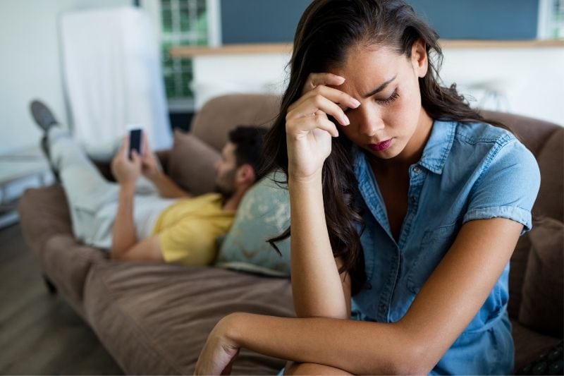 sad woman sitting on the couch with a man lying busy with his smartphone