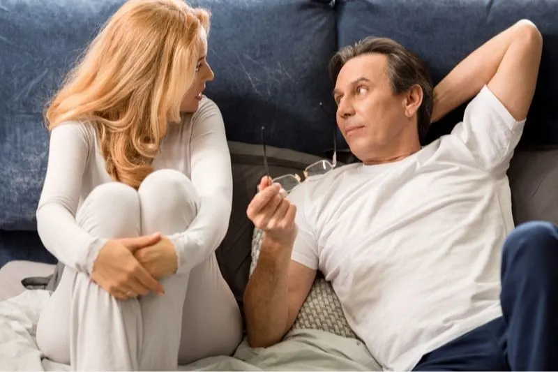 serious couple talking in bed with man lying holding an eyeglasses and woman sitting