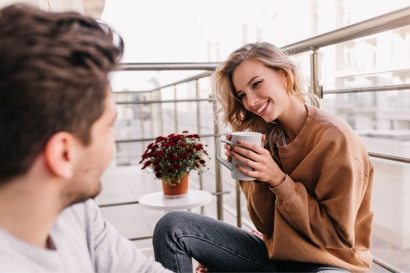 mujer tímida sonriendo mientras bebe de una taza y habla con un chico en la terraza