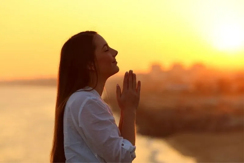 sideview of a praying woman with praying hands standing outdoors during golden hour