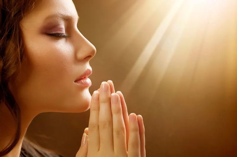 sidevoew of a pretty woman praying with hands close together with sun rays above
