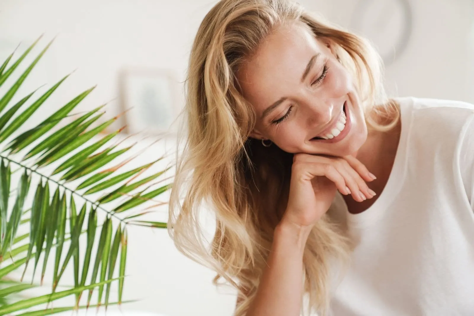 smiling woman closing her eyes sitting inside the living room with plant behind her