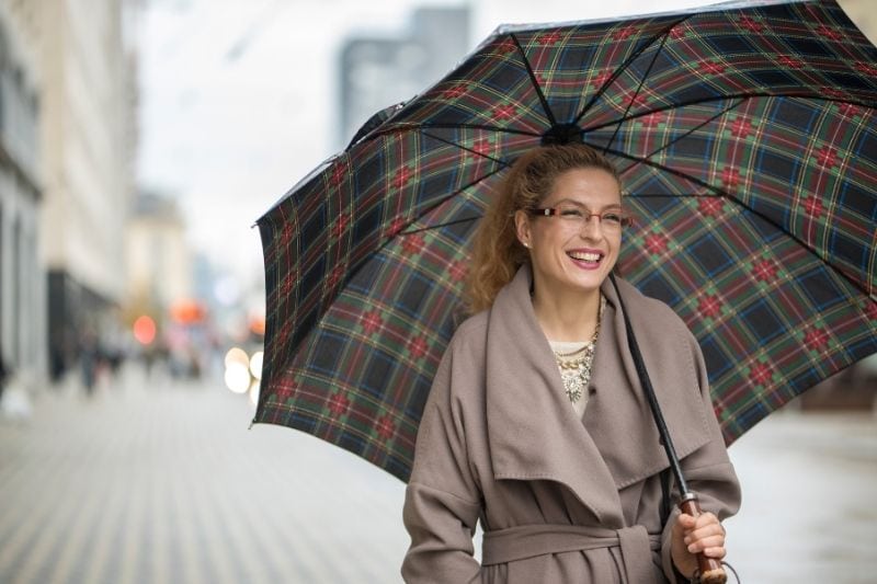 mulher sorridente com um guarda-chuva a descer a rua com uma gabardina comprida