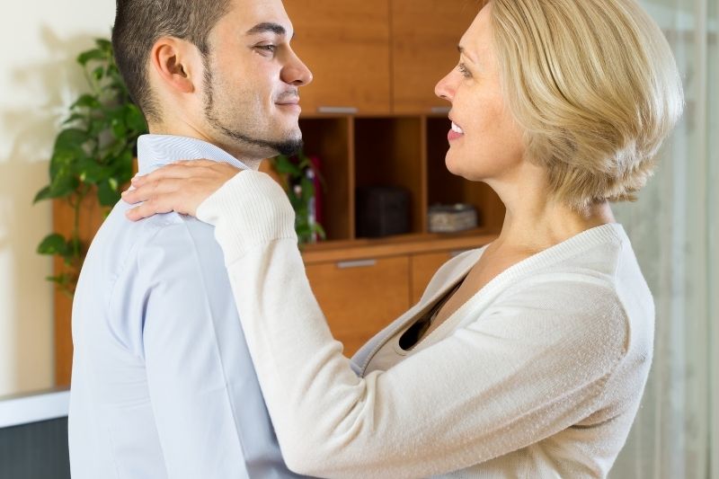 sweet mature woman slow dancing with a younger man inside living room.