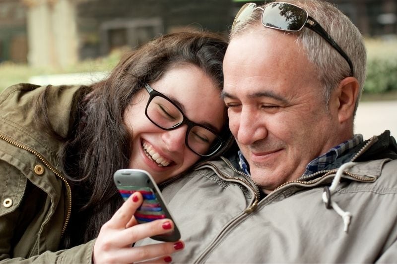teenage daughter leaning on her father's shoulder showing her phone smiling